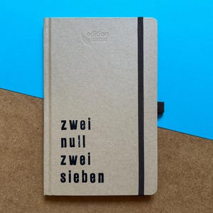 May include: A tan hardcover notebook with a black elastic closure band and a pen loop. The cover features the embossed text edition natur and the printed black text zwei null zwei sieben. The notebook has a textured, matte paper finish.