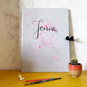 May include: A pale grey journal with the name "Jonna" in cursive, decorated with pink and navy blue paint splatters. A small black bow is attached to the side. A small cactus in an orange and beige pot sits nearby, along with pencils and a gold binder clip.