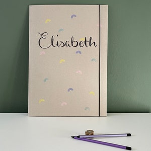 May include: A beige folder with a black elastic closure and the name "Elisabeth" written in black cursive. The folder is decorated with a pattern of small, colorful rainbows.