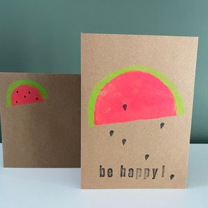 May include: A brown card with a pink watermelon slice with green rind and black seeds. The card says "be happy!" in black lettering.