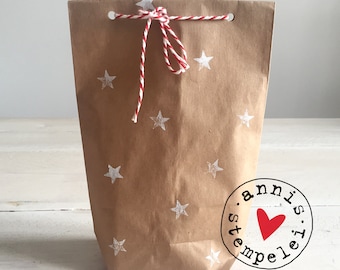 May include: A brown paper gift bag with white stars printed on it. The bag is tied with a red and white twine bow. A red heart with the text "annis.stempel.is." is printed on the bottom of the bag.