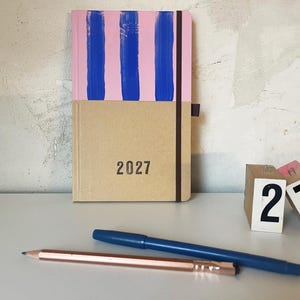 Könnte beinhalten: Ein rechteckiges Notizbuch mit einem rosa-blau gestreiften oberen Teil und einem beigen unteren Teil, auf dem das Jahr 2027 in Schwarz aufgedruckt ist. Ein dunkelbraunes Gummiband hält den Einband. Ein Metallstift und ein blauer Stift liegen davor.