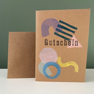 May include: A brown card with a geometric design in shades of pink, blue, and yellow. The word "Gutschein" is printed in black on the card.