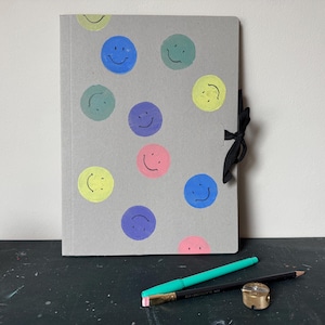 Könnte beinhalten: Ein graues Notizbuch mit bunten Smiley-Gesichtern. Das Notizbuch ist mit einem schwarzen Band gebunden und liegt auf einer dunklen Oberfläche neben einem türkisfarbenen Stift, einem Bleistift und einem Spitzer. Die Smileys sind in verschiedenen Farben.
