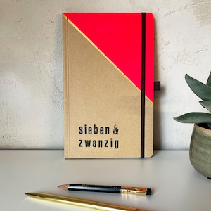 Könnte beinhalten: Ein rechteckiges Notizbuch mit einem beigen Einband, der einen diagonalen neonpinken Bereich und eine dünne goldene Linie aufweist. Der Text "sieben & zwanzig" ist in Schwarz auf dem unteren beigen Teil gedruckt. Ein schwarzes Gummiband hält das Notizbuch, das neben einem schwarzen Bleistift und einem goldenen Stift liegt.