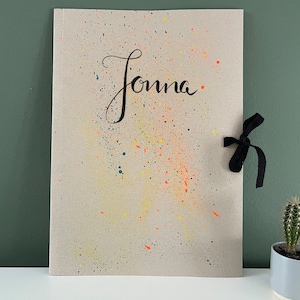 Puede incluir: Una carpeta de papel marrón con cierre de cinta negra. La carpeta tiene un patrón de salpicaduras de pintura amarilla, naranja y azul. El nombre "Jonna" está escrito en caligrafía negra en la parte delantera.
