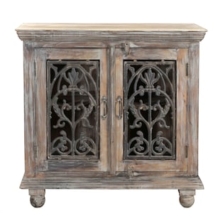 May include: A distressed wooden cabinet with two doors and ornate metal grillwork. The cabinet has a light-coloured top and is supported by four rounded feet. The doors have metal handles and a latch. The wood has a weathered, grey and brown finish.