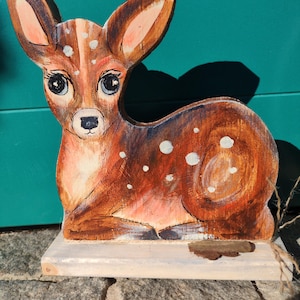 May include: A hand-painted wooden deer figurine in a resting position, featuring brown fur with white spots, large dark eyes, and long ears. The deer is mounted on a rectangular light-coloured wooden base with a small piece of bark attached.