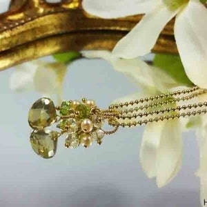 May include: A gold chain necklace with a green gemstone pendant. The pendant is surrounded by smaller green and white beads. The necklace is lying on a gold-colored surface.