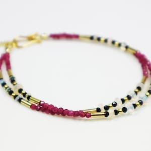 Könnte beinhalten: Doppelreihiges Armband mit kleinen, facettierten Rubinperlen, abwechselnd mit goldfarbenen zylindrischen Perlen und kleineren schwarzen und weißen Perlen. Das Armband hat einen goldfarbenen Verschluss.