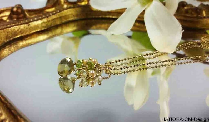 May include: A gold chain necklace with a green gemstone pendant. The pendant is surrounded by smaller green and white beads. The necklace is lying on a gold-colored surface.