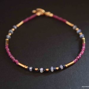 Könnte beinhalten: Zartes Armband mit kleinen, facettierten Rubinperlen, abwechselnd mit goldfarbenen zylindrischen Perlen und kleinen blauen und weißen Edelsteinen. Das Armband ist auf einem dunkelvioletten Band aufgezogen und hat einen goldenen Verschluss.