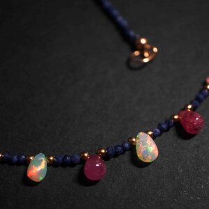 May include: A delicate necklace with a blue beaded chain and teardrop-shaped gemstones. The gemstones are a mix of pink and iridescent white.