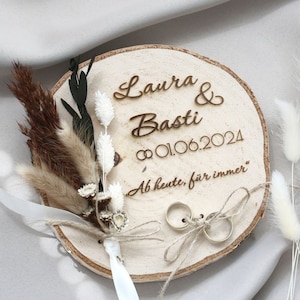 May include: Wooden ring bearer box with a rustic wood slice base. The box is decorated with dried flowers and a personalized inscription that reads "Laura & Basti 01.06.2024 Ab heute, für immer".