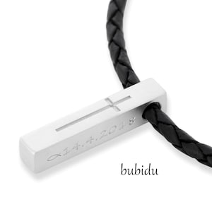 May include: A silver cross pendant with the date "11.4.2018" engraved on it, attached to a black braided leather cord.
