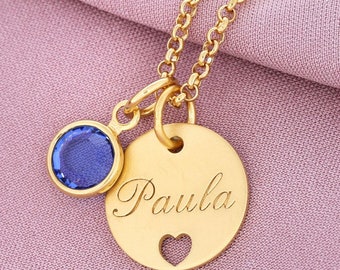 Puede incluir: Collar de oro con un colgante redondo que dice "Paula" con un pequeño corazón recortado. El colgante está unido a una pequeña piedra preciosa azul.
