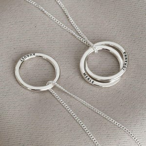 Könnte beinhalten: Zwei silberne Halsketten mit ineinandergreifenden Ringen. Der größere Ring trägt die Namen "Merle" und "Johanna" eingraviert. Der kleinere Ring trägt den Namen "August" eingraviert.