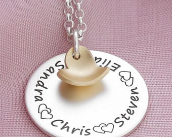 Puede incluir: Collar de plata con un colgante redondo con los nombres "Chris", "Steven", "Ellie" y "Sandra" grabados alrededor del borde, con corazones entre cada nombre. Un dije de corazón dorado cuelga del colgante.