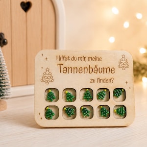 Könnte beinhalten: Ein rechteckiges helles Holzbrett mit dem eingravierten deutschen Text 'Hilfst du mir, meine Tannenbäume zu finden?' und zehn quadratischen Aussparungen, die jeweils eine kleine grüne Kunststoff-Tannenbaumfigur mit gelbem Stern enthalten.
