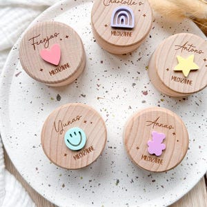 Könnte beinhalten: Fünf kleine, runde Holzkästchen mit personalisierten Namen und dekorativen Aufsätzen, darunter ein Herz, ein Regenbogen, ein Stern, ein Smiley und ein Teddybär. Auf jedem Kästchen ist der Name "MILCHZAHNE" eingraviert. Die Kästchen liegen auf einem gesprenkelten weißen Teller.