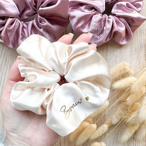 Könnte beinhalten: Ein Set aus drei Satin-Haargummis in den Farben Pink und Creme. Die Haargummis sind mit goldenen Buchstaben verziert. Das cremefarbene Haargummi sagt "Bride" und das rosafarbene Haargummi sagt "Team Bride".