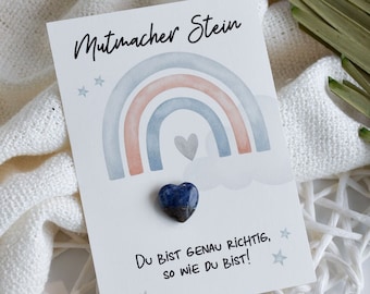 May include: A card with a rainbow design and the text "Mutmacher Stein" and "Du bist genau richtig, so wie du bist!" A blue heart-shaped stone is attached to the card.