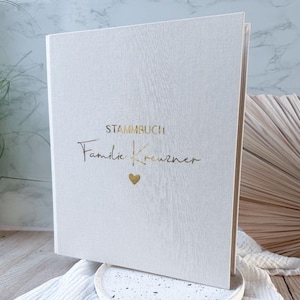 Könnte beinhalten: Ein weißes Stammbuch, oder Familienbuch, mit goldfarbenen Buchstaben und einem kleinen goldenen Herz. Das Buch steht auf einem weißen und gesprenkelten runden Tablett. Der Text auf dem Cover lautet "STAMMBUCH" und "Familie Kreuzner".