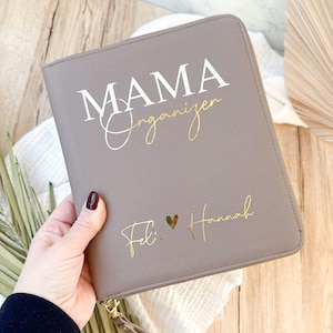 Könnte beinhalten: Ein taupefarbener Organizer mit den Worten "MAMA Organizer" in weißer und goldener Schrift. Die Namen "Feli" und "Hannah" sind in goldener Schrift darunter gedruckt, mit einem goldenen Herzsymbol dazwischen. Der Organizer hat einen Reißverschluss und eine Handschlaufe.