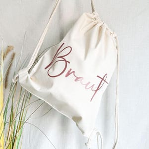 Könnte beinhalten: Eine weiße Kordeltasche mit dem Wort "Braut" in roségoldener Schrift.
