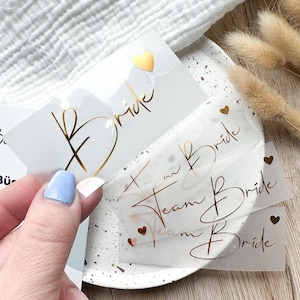 Könnte beinhalten: Vier transparente Aufkleber mit goldener Folienbeschriftung. Die Aufkleber sagen "Bride", "Team Bride", "Team Bride" und "Bride".