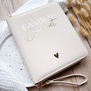 Könnte beinhalten: Eine cremefarbene Reißverschluss-Clutch mit dem Schriftzug "FAMILIE Gebhardt" in weißer und goldener Schrift. Ein kleines goldenes Herz befindet sich unten rechts. Die Clutch hat eine Handschlaufe und goldfarbene Hardware.