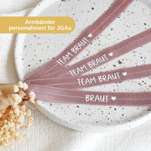 Könnte beinhalten: Vier rosafarbene elastische Armbänder mit weißem Text, der "Team Braut" und "Braut" mit einem kleinen Herzsymbol lautet. Die Armbänder liegen auf einer weißen, gesprenkelten Oberfläche.