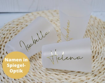 May include: Three frosted glass cups with gold lettering. The names "Isabella", "Helena", and "Andrea" are written in a script font on the cups. The text "Namen in Spiegel-Optik" is written in black on a yellow background.