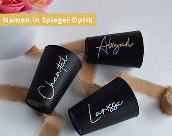 May include: Three black tumblers with iridescent lettering spelling out the names Abigail, Chantal, and Larissa. The tumblers are arranged on a white surface with a tan ribbon and a white vase with pink and orange flowers in the background. The text "Namen in Spiegel-Optik" is in a yellow box.