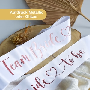 Könnte beinhalten: Zwei weiße Satinschleifen mit roségoldenen Schriftzügen. Eine Schleife sagt "Team Bride" und die andere "Bride to be" mit einem Herzsymbol.