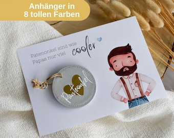May include: A gray fabric keychain with a gold heart and the text "Patenonkel" in gold lettering. The keychain is attached to a gold metal ring and a gold metal clasp. The keychain is displayed on a white card with the text "Patenonkel sind wie Papas nur viel cooler" in black lettering.