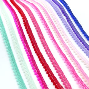 Puede incluir: Una colección de cintas de adorno con pompones coloridos en varios tonos de rosa, rojo, morado, azul y verde. Las cintas están dispuestas en diagonal, mostrando sus texturas y colores. Los pompones son pequeños y espaciados.