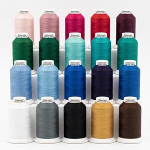 Op de afbeelding: Een verzameling spoelen Gutermann Minking 1000 garen in diverse kleuren, waaronder roze, rood, groen, blauw en zwart. Elke spoel is voorzien van het merk en het type garen. De spoelen zijn gerangschikt op een witte display.