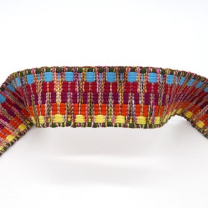 Peut inclure: Une garniture tiss&eacute;e color&eacute;e avec un motif r&eacute;p&eacute;titif de rectangles rouges, oranges, jaunes, bleus et verts. La garniture est incurv&eacute;e, montrant sa flexibilit&eacute;. Le fond est blanc uni.