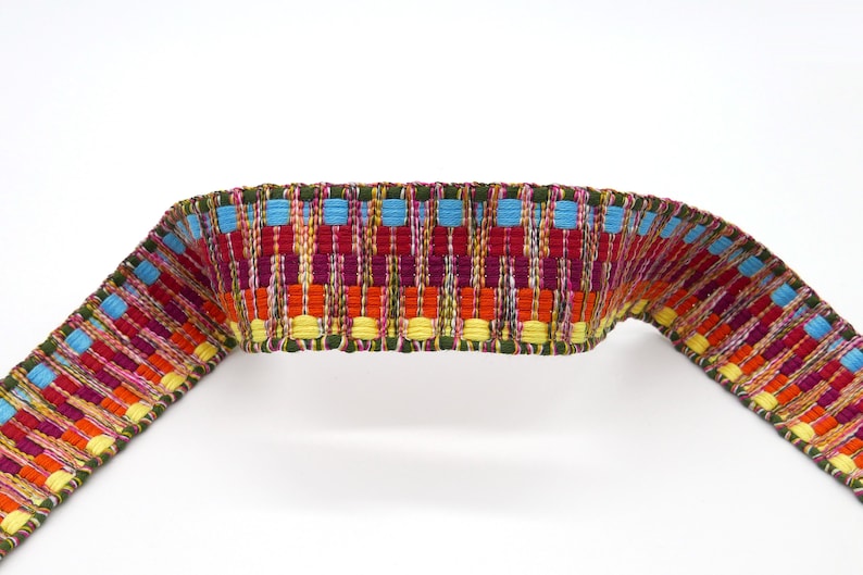 Peut inclure: Une garniture tiss&eacute;e color&eacute;e avec un motif r&eacute;p&eacute;titif de rectangles rouges, oranges, jaunes, bleus et verts. La garniture est incurv&eacute;e, montrant sa flexibilit&eacute;. Le fond est blanc uni.