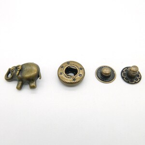Pode incluir: Um conjunto de quatro botões de metal de cor dourada antiga. Um botão apresenta um design de elefante, enquanto os outros três são redondos com um padrão de estrelas. Os botões redondos têm um furo no centro para costura.