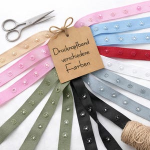May include: A selection of colourful snap tape ribbons in shades like pink, blue, and green. A brown tag with the text "Druckknopfband verschiedene Farben" is attached. Scissors and a ball of twine are also visible.