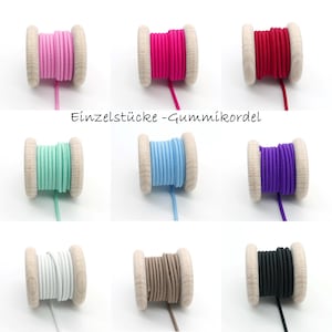 Puede incluir: Nueve carretes de cordón elástico de colores, rosa, fucsia, rojo, verde menta, azul claro, morado, blanco, marrón y negro. Cada carrete está enrollado alrededor de un núcleo de madera. El texto "Einzelstücke - Gummikordel" está impreso en el centro.