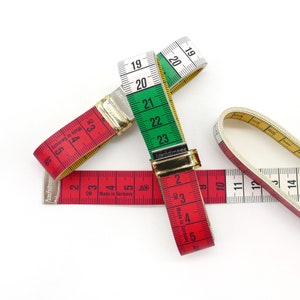 Può includere: Tre nastri per misurare, uno rosso e bianco, uno verde e bianco e uno bianco con numeri neri. Il nastro rosso e bianco riporta "CM Made in Germany" e il nastro verde e bianco riporta "Made in Germany".