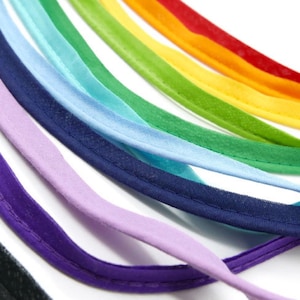 Op de afbeelding: Een regenboogassortiment van kleurrijk stoffenbies met een gestikte rand. De bies is in een halve cirkel gerangschikt, met de rode bies bovenaan en de paarse bies onderaan.