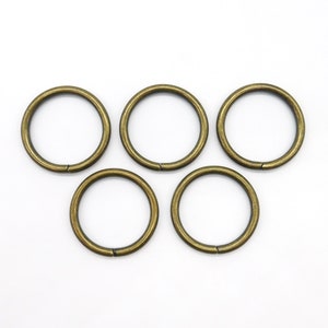 Könnte beinhalten: Fünf bronzefarbene Metallringe auf weißem Hintergrund. Die kreisförmigen Ringe haben eine kleine Lücke, was darauf hindeutet, dass sie geöffnet und geschlossen werden können. Sie haben ein leicht gealtertes, Vintage-Aussehen.