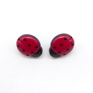 Peut inclure: Une paire de boucles d'oreilles à clous en forme de coccinelles. Chaque boucle d'oreille présente un corps rouge brillant avec des points noirs et une tête noire. Les boucles d'oreilles sont sur fond blanc.