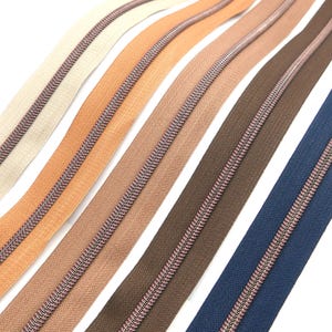 Könnte beinhalten: Sortiment von Reißverschlussbändern in verschiedenen Farben, darunter Beige, Braun, Orange und Blau. Die Reißverschlüsse haben einen silbernen Metall-Schieber und einen rosafarbenen Faden, der entlang der Zähne verläuft.