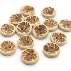 May include: A collection of round wooden buttons with a light tan base and a brown leaf design. Each button has two holes for sewing. The buttons are scattered on a white surface.