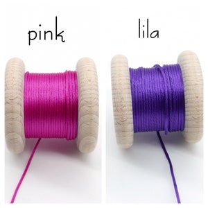 Peut inclure: Deux bobines en bois enroul&eacute;es de cordon. La bobine de gauche pr&eacute;sente un cordon rose vif avec le texte 'pink' au-dessus. La bobine de droite pr&eacute;sente un cordon violet fonc&eacute; avec le texte 'lila' au-dessus. Les deux bobines ont une finition en bois clair.